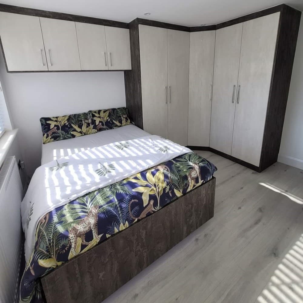 Fitted corner wardrobe unit with two-tone upper cabinets and drawers, installed above and around a bed in a bespoke bedroom layout.