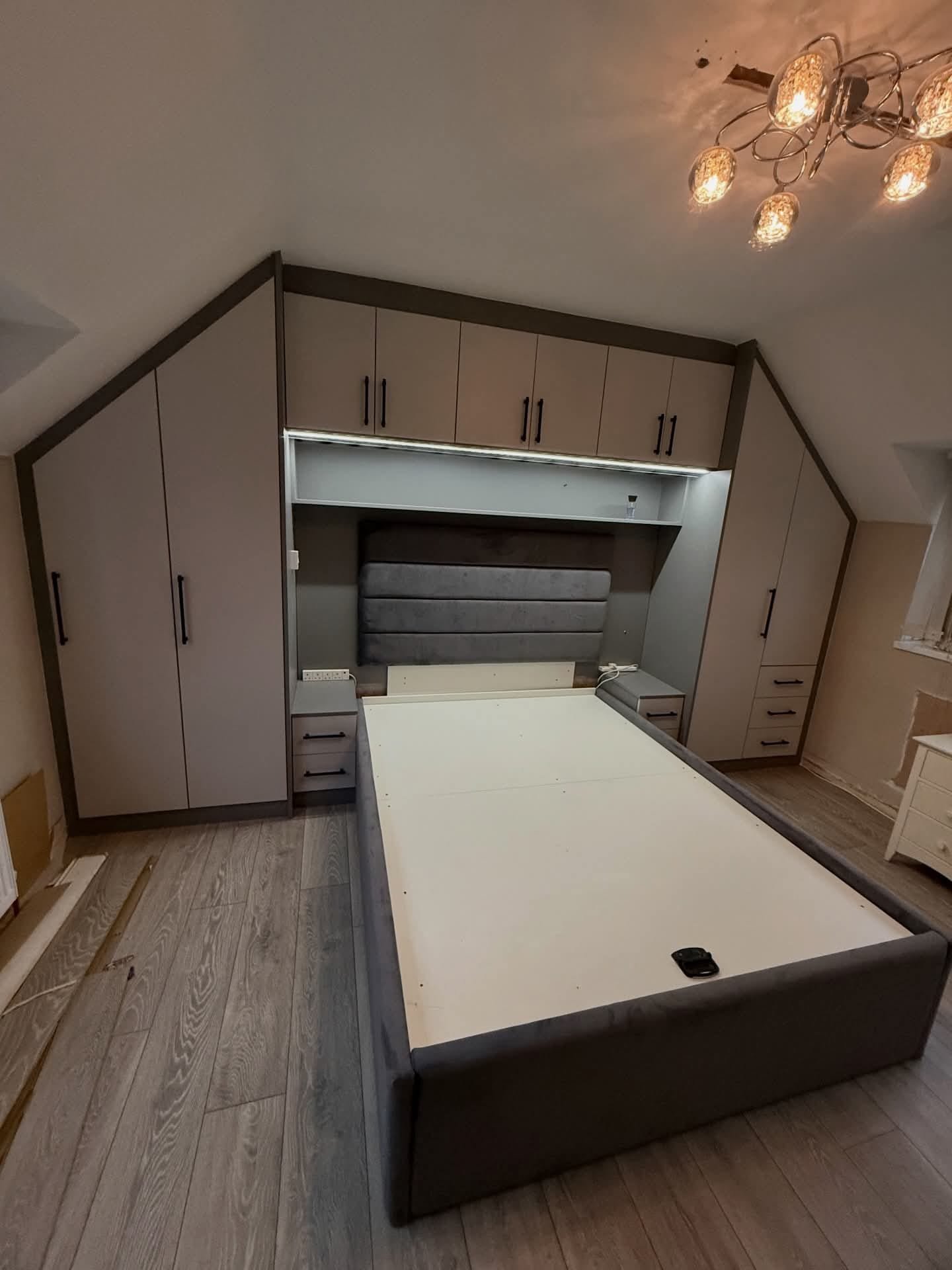 Eaves storage solution with custom fitted wardrobes, over-bed cabinet unit and LED lighting in loft room with angled ceiling — taupe finish with dark brown trim